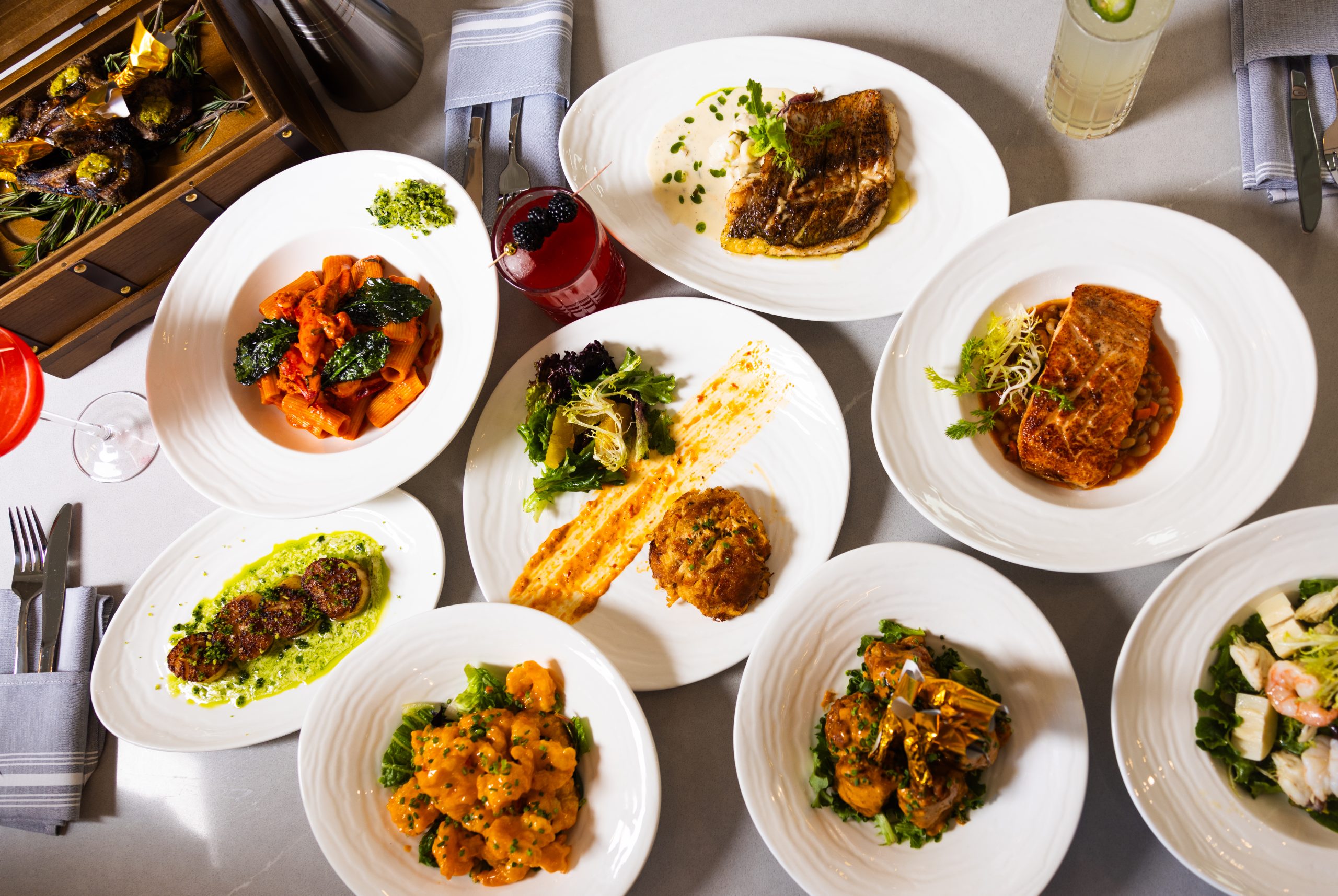 A beautifully arranged spread of OU LA restaurant’s signature dishes, including grilled lamb chops served in a wooden treasure chest, creamy seafood pasta, Caribbean-spiced salmon, seared scallops, and flavorful crab cakes. The vibrant plating and fine dining presentation highlight the restaurant’s fusion of global techniques with Caribbean influences, accompanied by handcrafted cocktails.