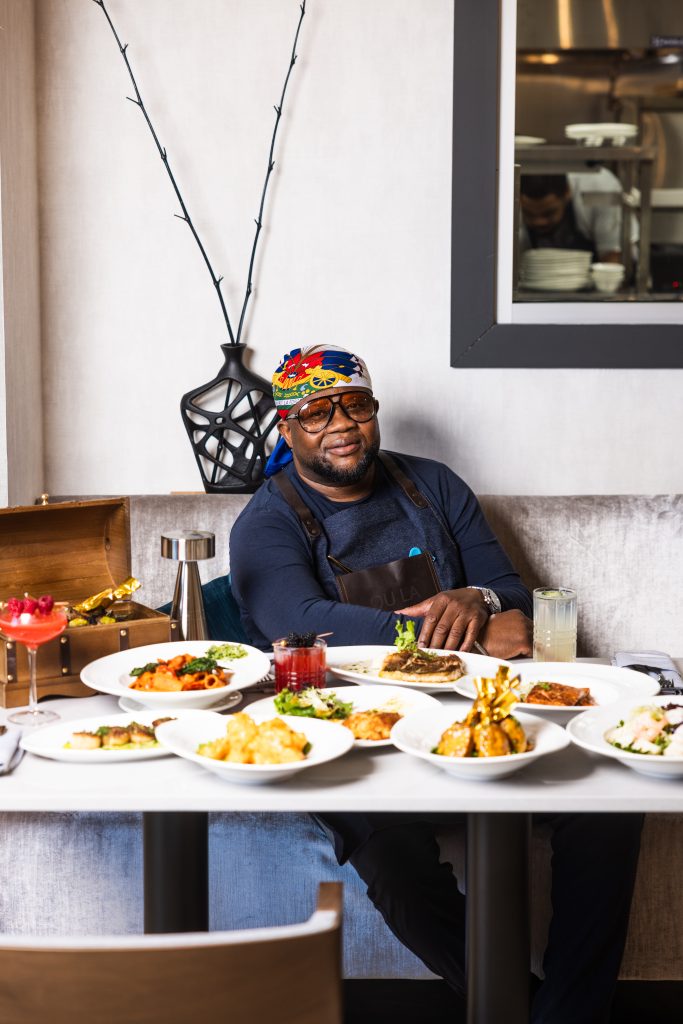 Chef Duke of OU LA restaurant, wearing a Haitian flag bandana and stylish sunglasses, sits proudly at a table adorned with an array of his signature Caribbean fusion dishes. The spread includes grilled lamb chops in a wooden treasure chest, seafood pasta, seared scallops, crab cakes, and handcrafted cocktails. The open kitchen in the background adds to the dynamic dining experience at OU LA.