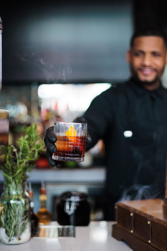 A bartender at OU LA restaurant presenting a smoked craft cocktail with an orange peel garnish. The rich, aromatic drink is served in an elegant glass, creating a sophisticated and immersive mixology experience at the upscale Caribbean-inspired bar.